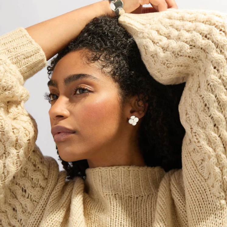 A woman wearing a chunky cable-knit beige sweater, posing with her arms raised against a light gray background.