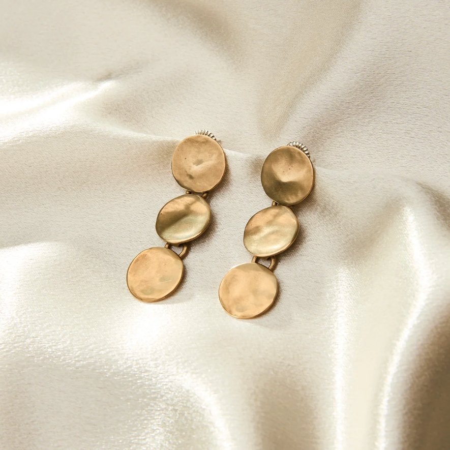 A pair of gold disc drop earrings with a hammered texture, displayed on a draped cream satin fabric background.