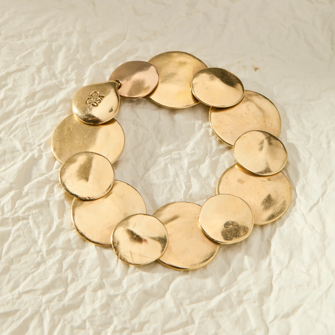 A gold-tone disc bracelet made of overlapping round coin-like discs, photographed on a textured off-white paper background.