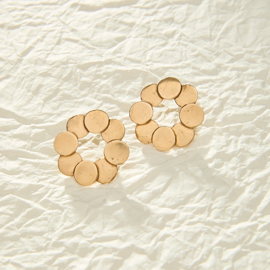 A pair of gold-toned flower-shaped stud earrings made of multiple small circular discs, resting on a textured, crumpled off-white paper background.