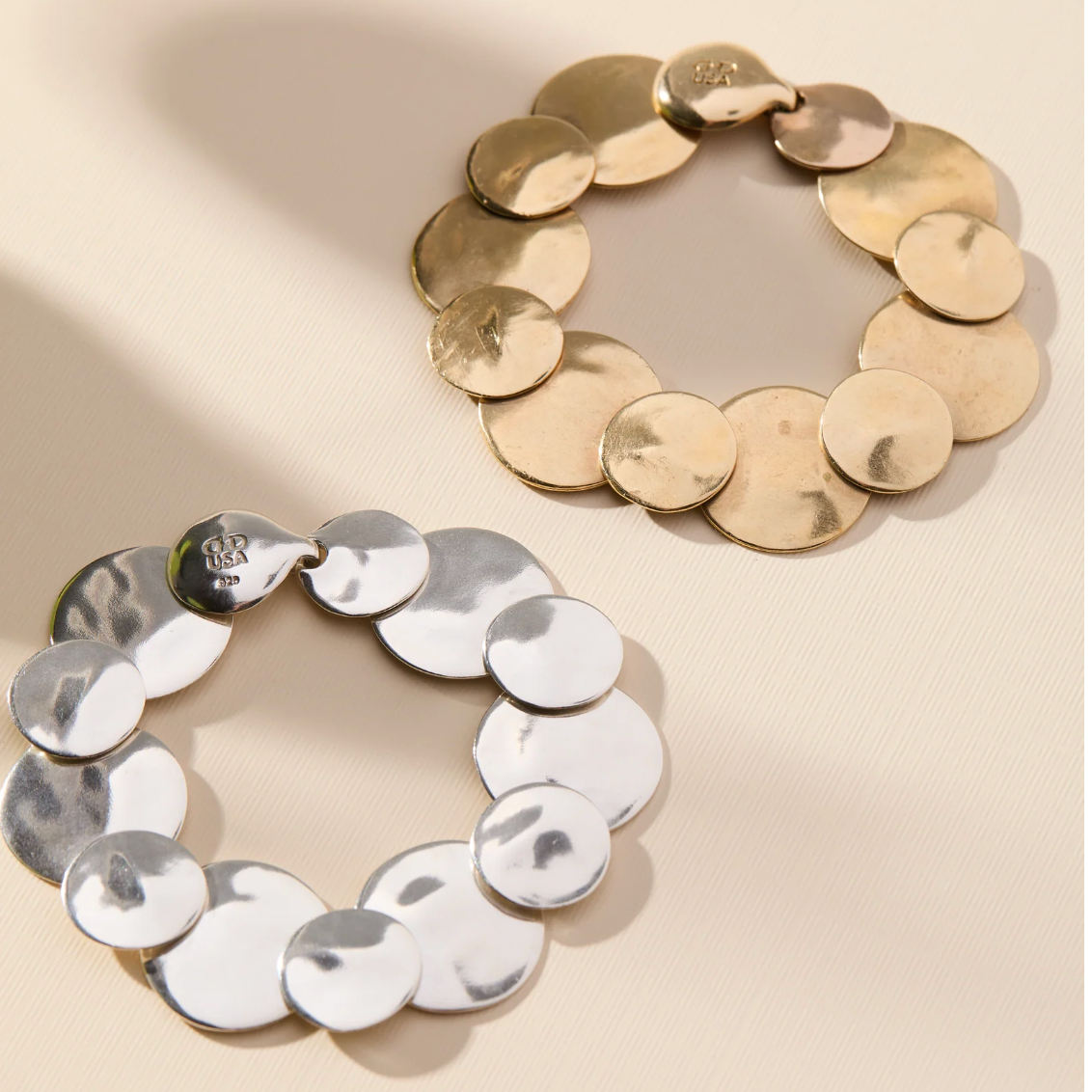 Two circular disc-link earrings, one in polished silver and one in polished gold, displayed on a warm beige background with soft shadows.