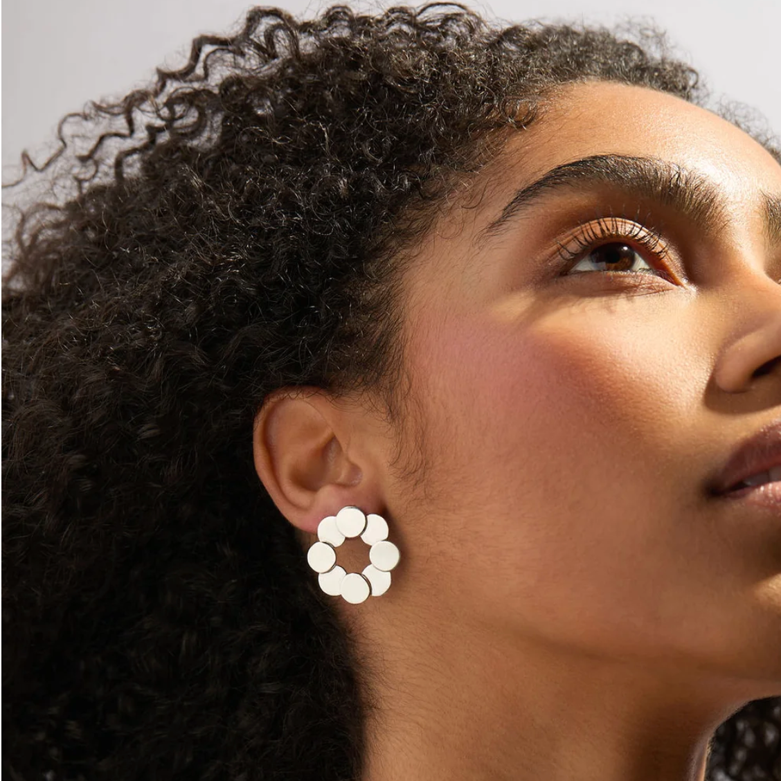 A close-up side profile of a woman with curly dark hair wearing a white flower-shaped stud earring with a silver-toned metal setting.