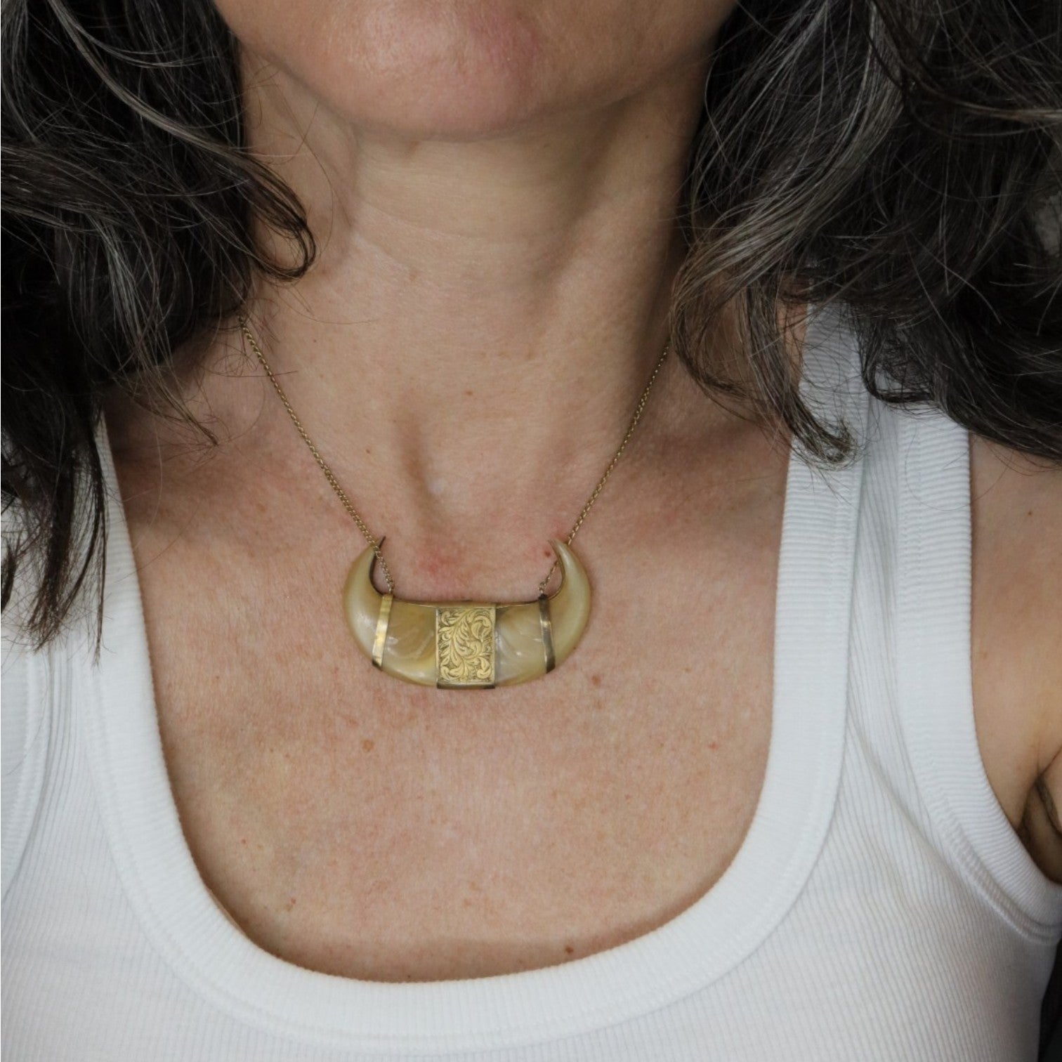 A close-up of a person wearing a gold crescent moon pendant necklace with a textured square center, styled with a white ribbed tank top.