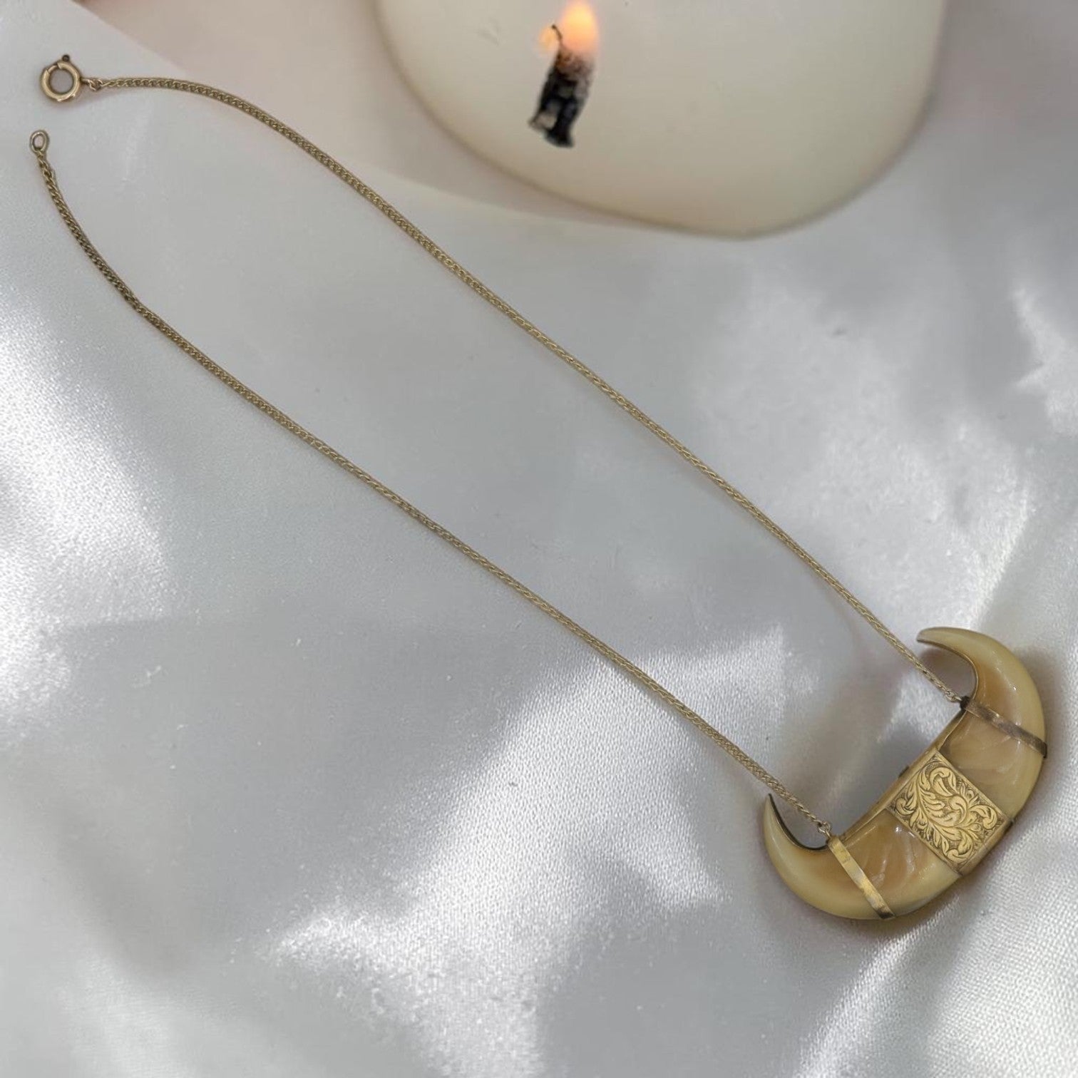A gold chain necklace with a crescent moon pendant featuring a carved beige bone or shell inlay, displayed on a white satin fabric next to a lit candle.