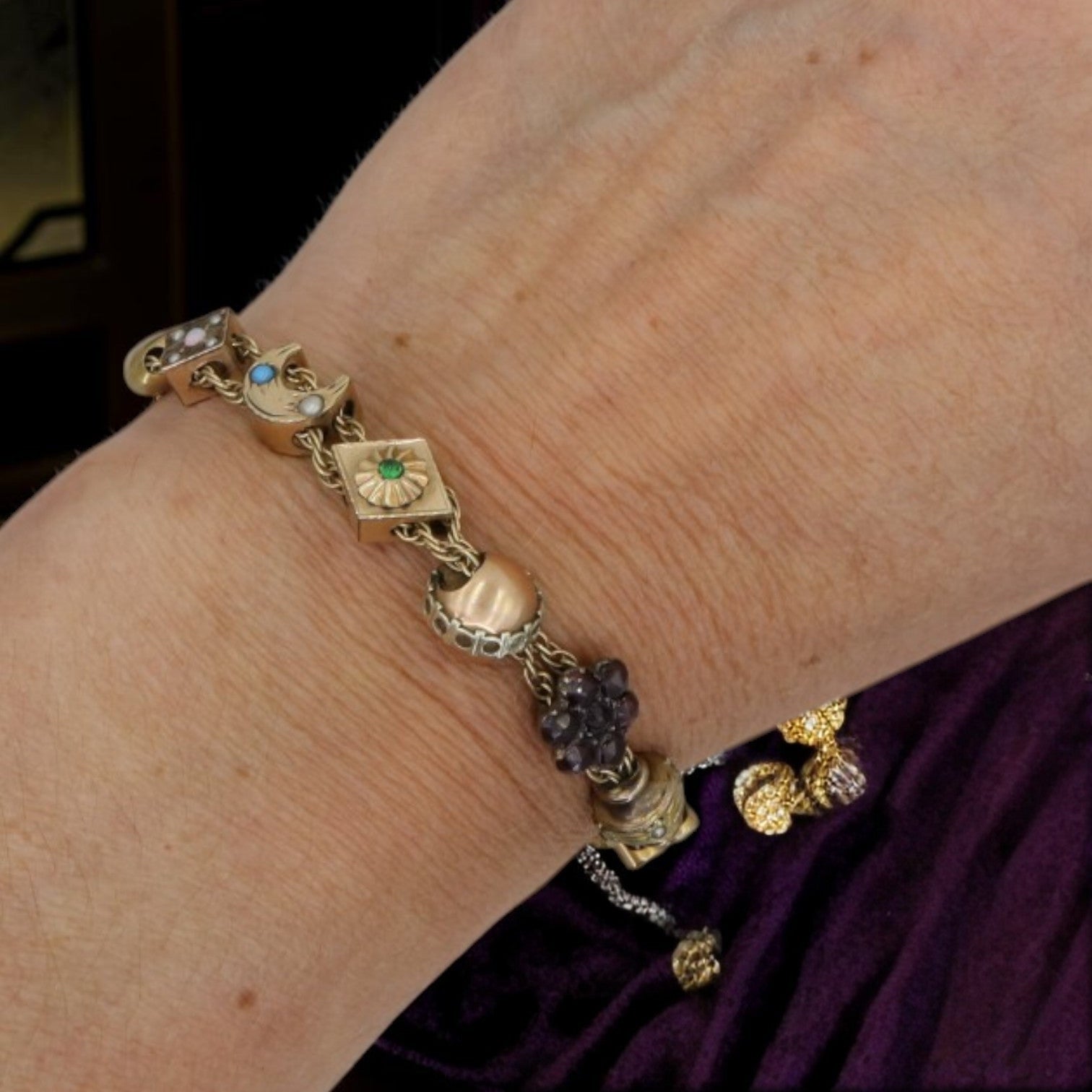 A close-up of a person's wrist wearing a gold charm bracelet with multiple decorative charms, including a green sun charm, a blue stone charm, and a dark purple grape cluster charm, set against a dark background.