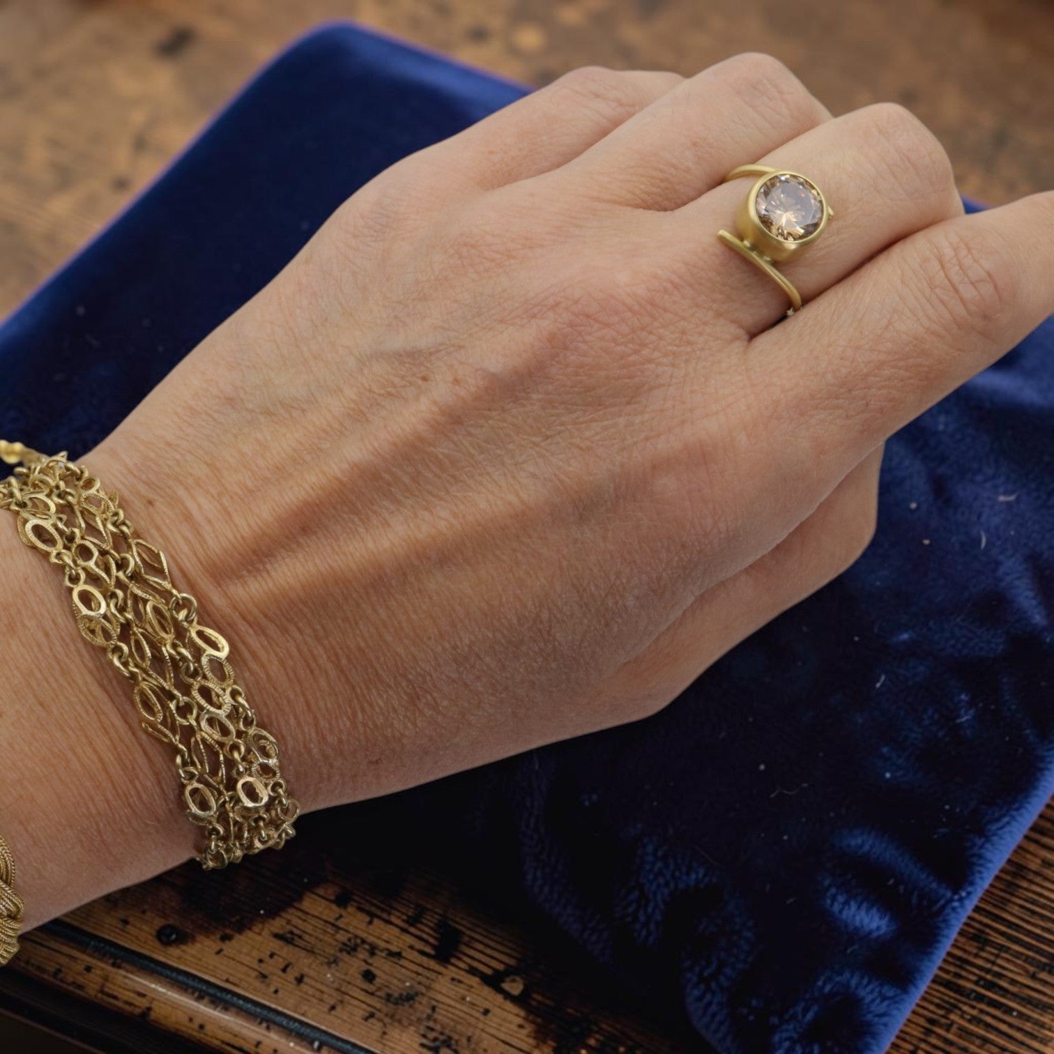 A close-up of a person's hand wearing a yellow gold ring with a round clear gemstone and a matching yellow gold link bracelet, resting on a dark blue velvet jewelry box.